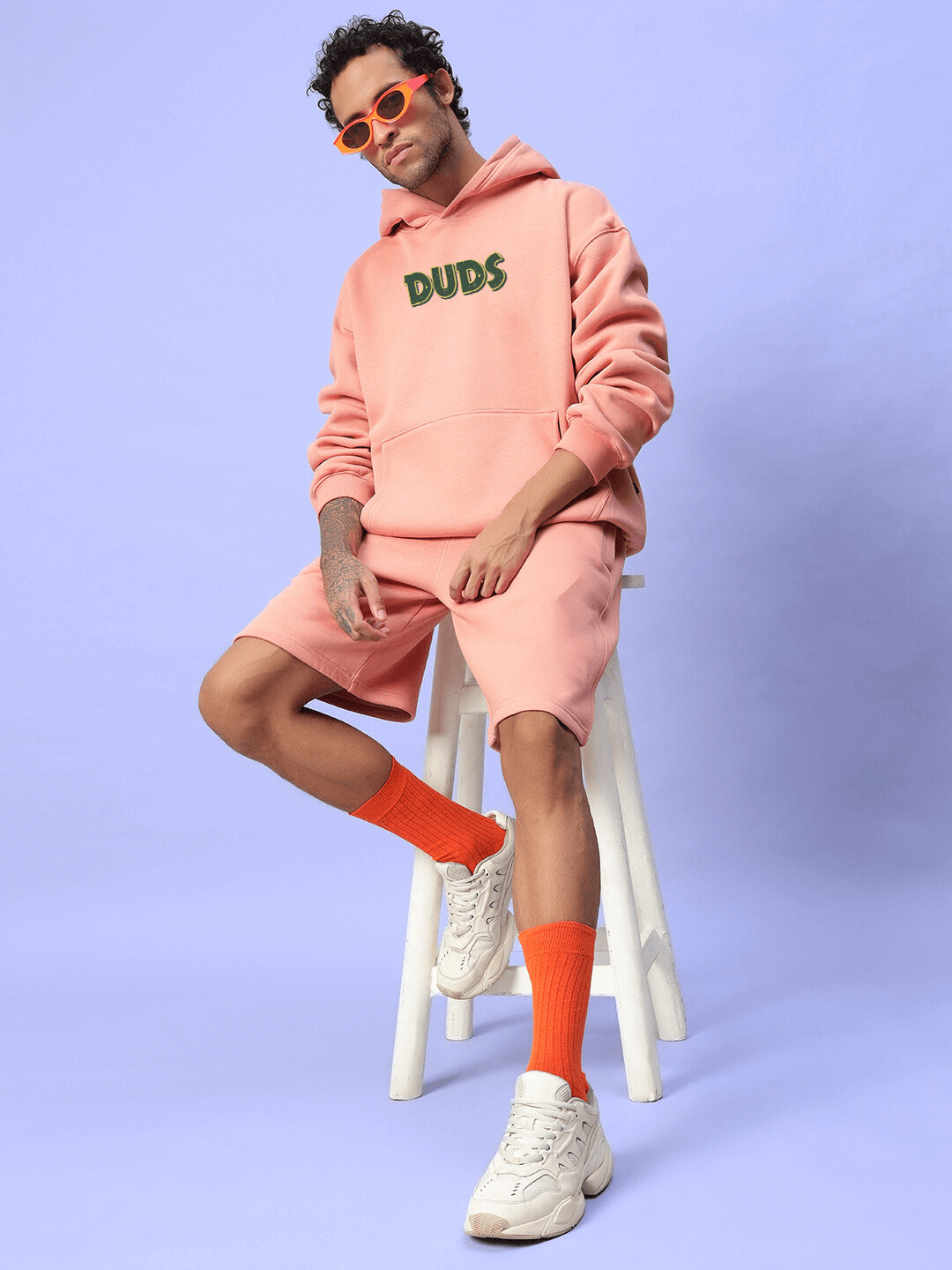 Man wearing JUDY NICK ZOOTOPIA FLEECE CO-ORD PEACH outfit with hoodie and shorts, sitting on a stool, showcasing casual streetwear style.