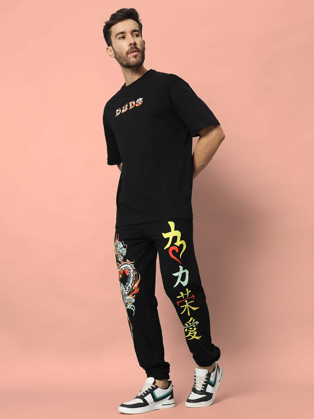 Man wearing KAKAAI Relaxed Pant Jogger in black with colorful designs, standing against a pink background. Fashion by DUDS.