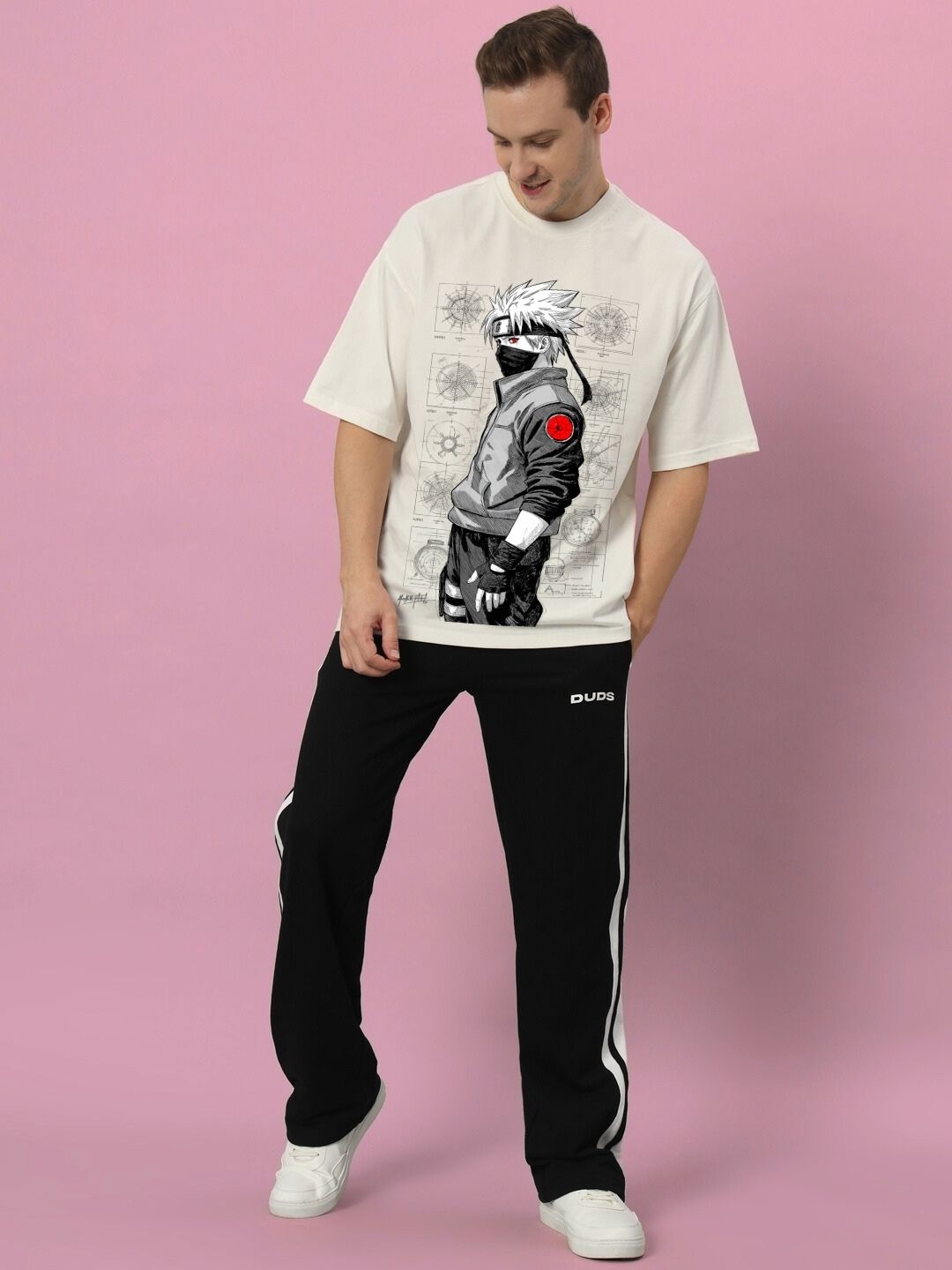 Man wearing KAKASHI PRINTED OVERSIZED T-SHIRT OFF-WHITE with anime character design, styled with black pants and white shoes, standing against pink background.