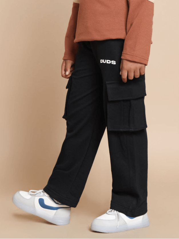 Child wearing black Keep Flying jogger pants for boys and girls with side pockets and DUDS logo, paired with white sneakers.