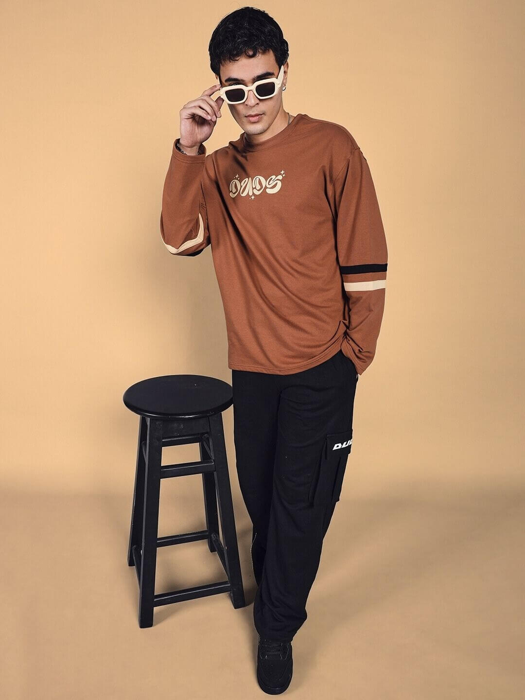 Model wearing brown oversized sweatshirt with black trousers, styled with white sunglasses, standing beside a black stool