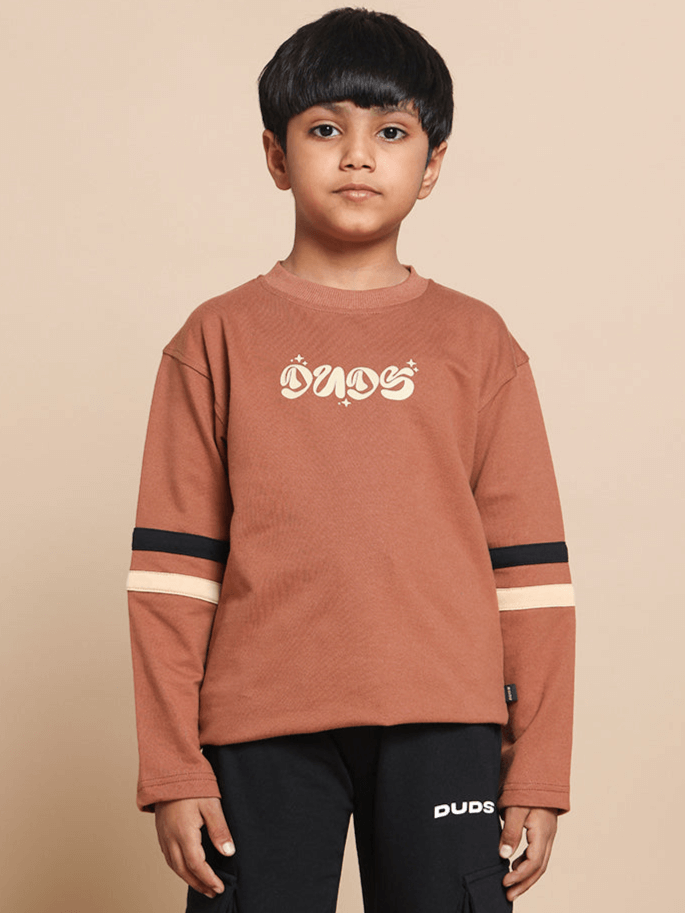 Young boy wearing a brown oversized sweatshirt with 'DUDS' printed on the front, featuring black stripe details on the sleeves.