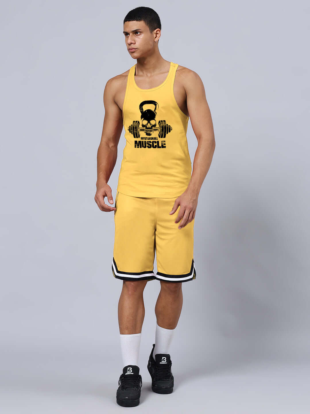 Man wearing a KETTLEBELL SKULL PRINTED GYM CO-ORD SET YELLOW, featuring a tank top and shorts in a fitness studio setting.