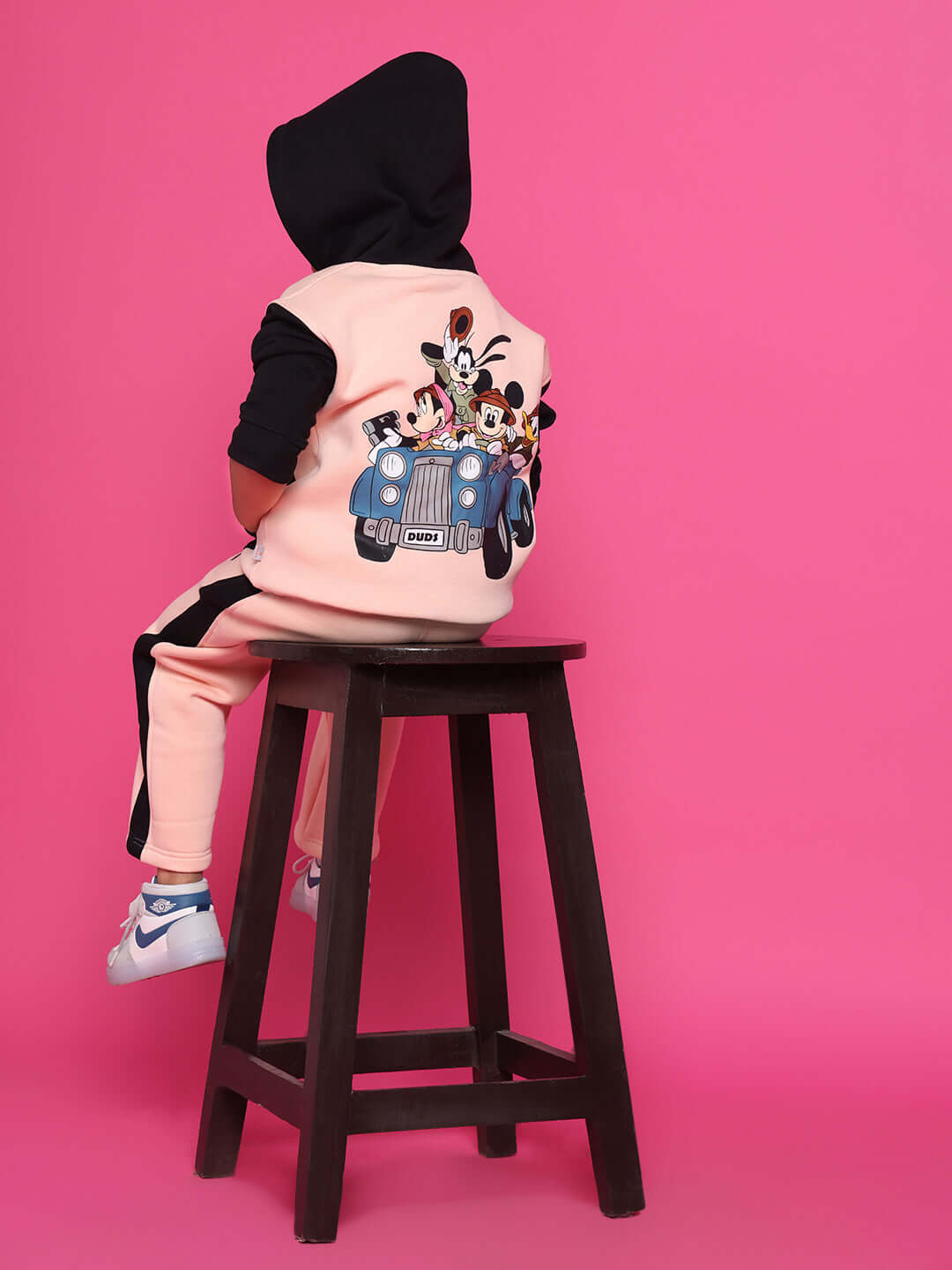 Child wearing a black and beige club co-ord set with colorful cartoon characters on the back, sitting on a stool against a pink background.