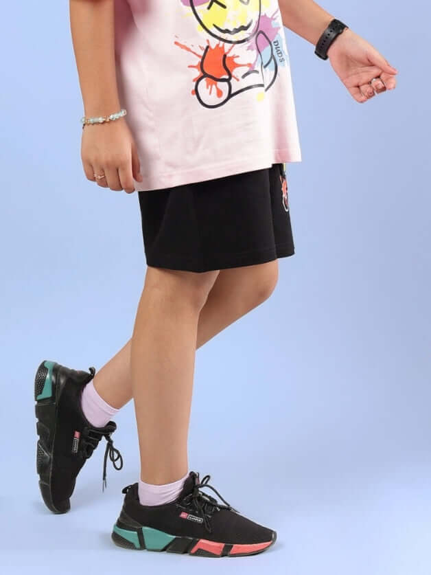 Child wearing BE KIND SHORTS FOR BOYS & GIRLS BLACK standing stylishly with colorful shoes on blue backdrop.