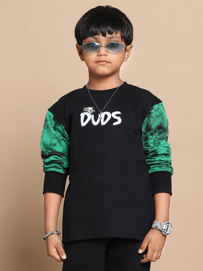 Boy wearing a black 'DUDS' oversized sweatshirt with green sleeves, standing against a beige background.