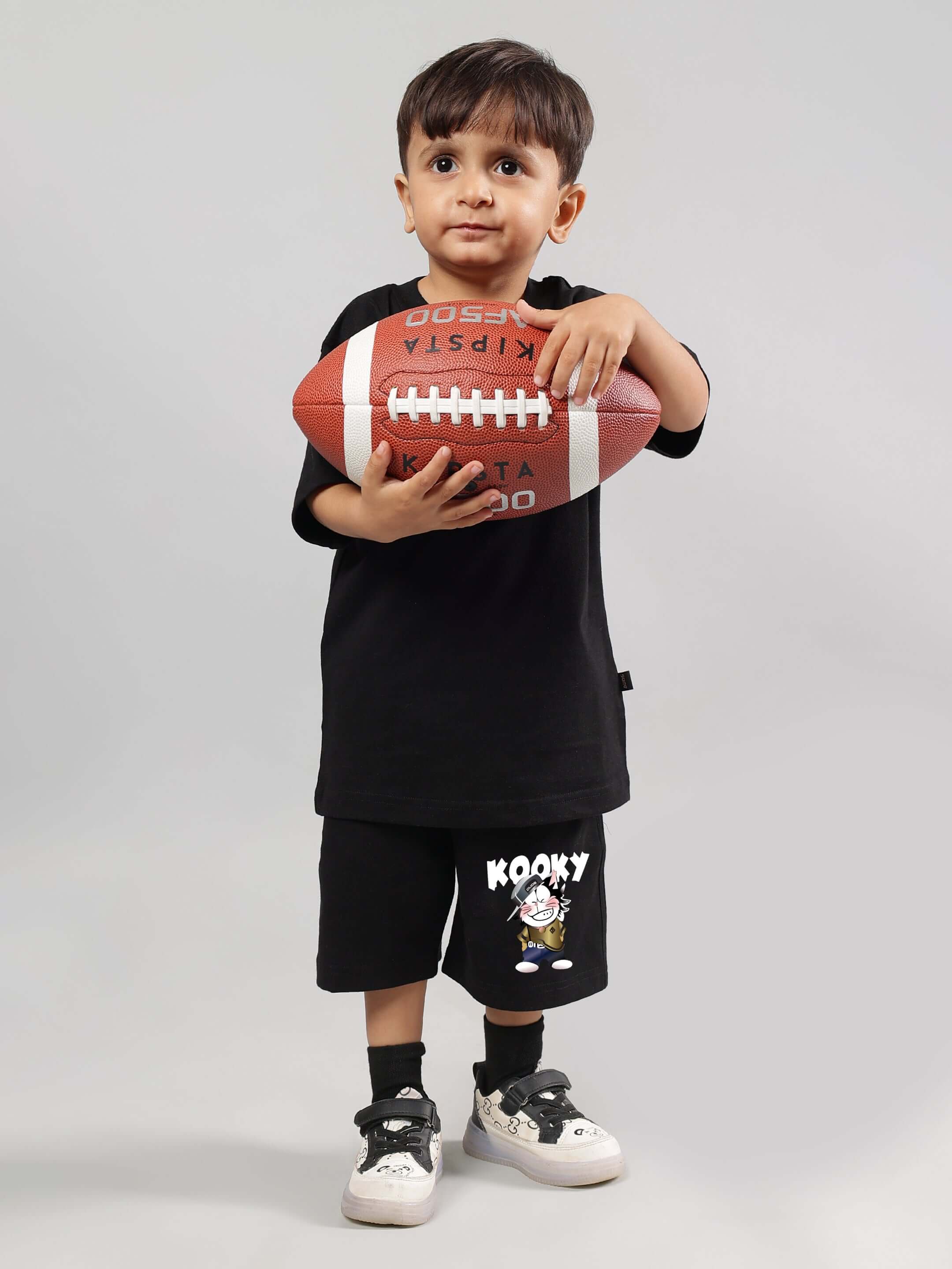 Young child wearing Kooky black shorts holding a football, showcasing customizable children's clothing by DUDS.