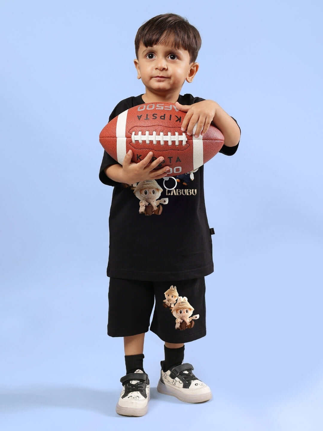 Young boy wearing a black LABUBU GANG T-shirt and shorts, holding a football.