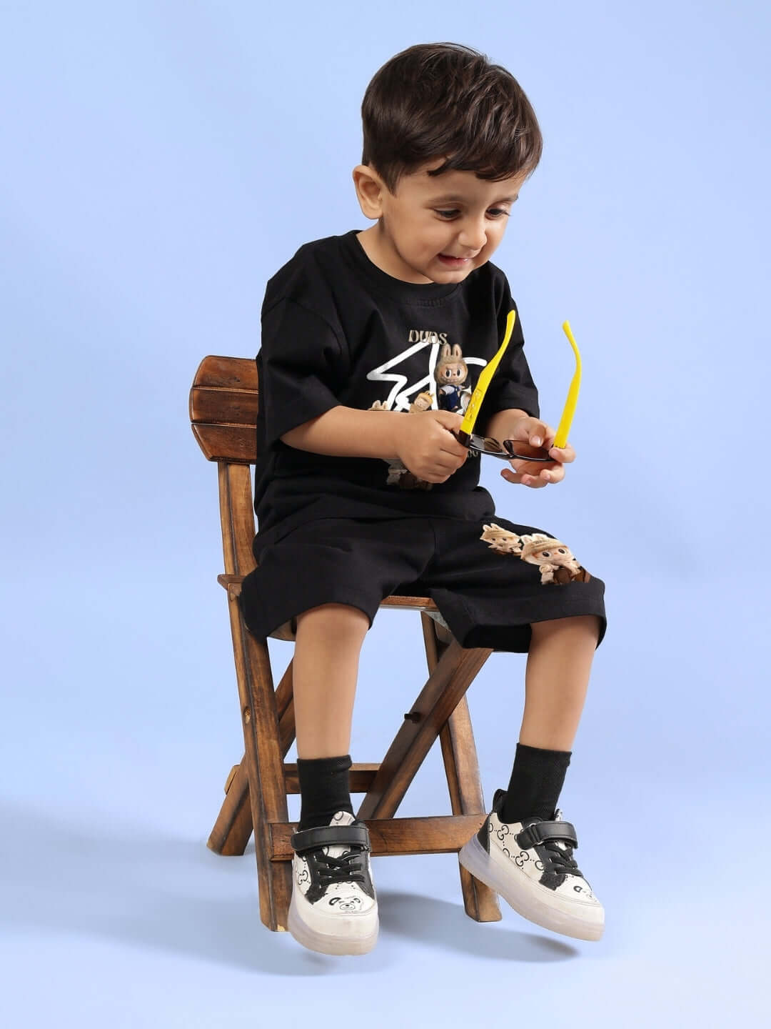Young child wearing a black LABUBU GANG T-shirt for boys and girls, sitting on a wooden chair, holding yellow glasses.