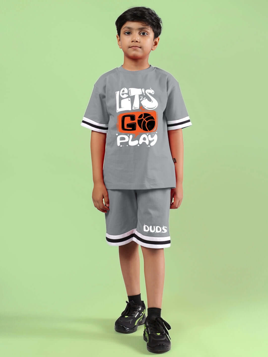 Child wearing grey "Let's Go Play" printed co-ord set for boys and girls by DUDS against a green background.