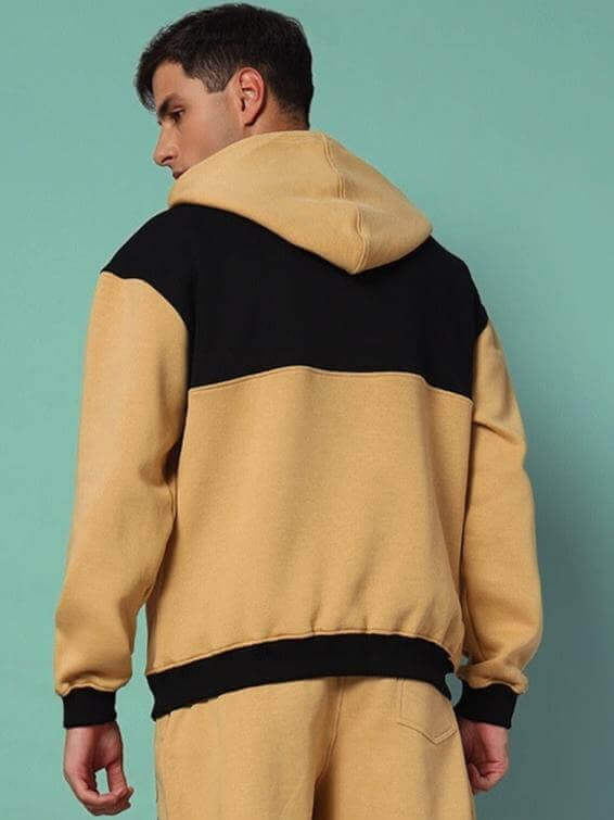 Man wearing LIBERTY FLEECE COLORBLOCK HOODIE BEIGE BLACK showcasing the stylish two-tone design against a teal background.