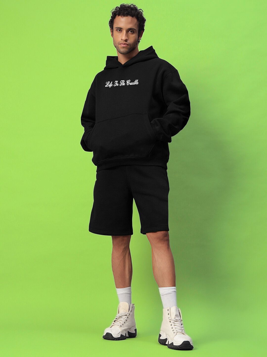 Model wearing LIFE IN THE CRUCIBLE PRINTED FLEECE CO-ORD BLACK, showcasing sleek and comfortable streetwear style on a green background.