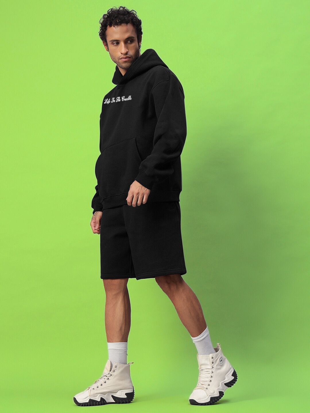 Model wearing LIFE IN THE CRUCIBLE PRINTED FLEECE CO-ORD BLACK against green background, showcasing streetwear style.