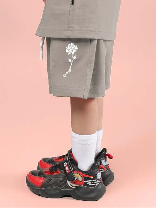 Child wearing LIMBO SHORTS FOR BOYS & GIRLS GREY with rose print, paired with colorful sneakers on a pink background.
