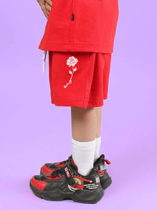 Child wearing LIMBO SHORTS FOR BOYS & GIRLS RED with floral embroidery, standing in stylish sneakers against a purple background.