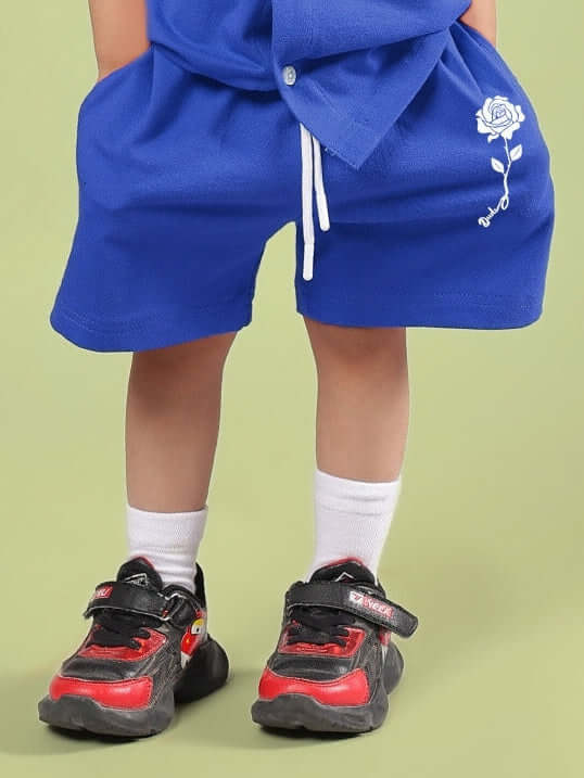 Child wearing LIMBO SHORTS FOR BOYS & GIRLS BLUE, showing front view with sneakers on a green background.