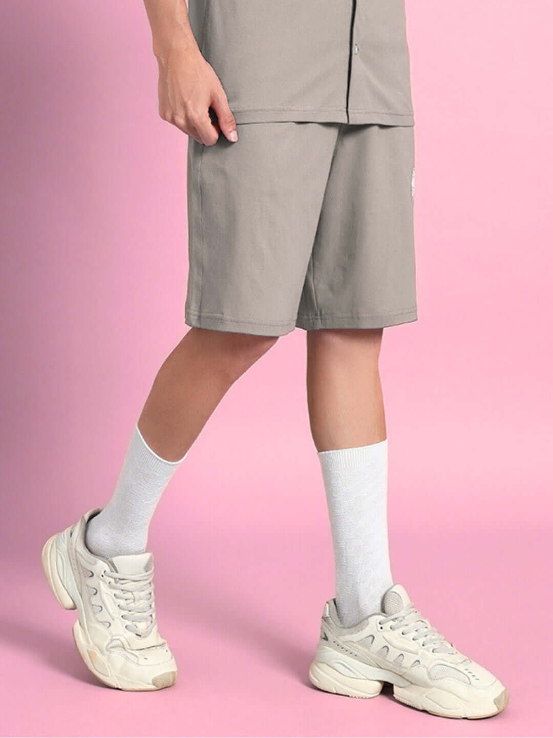 Person wearing LIMBO SUMMER COTTON SHORTS GREY with white socks and sneakers against a pink background.