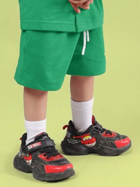 Child wearing LIMBO SHORTS FOR BOYS & GIRLS GREEN, standing in colorful shoes with white socks against a light green background.