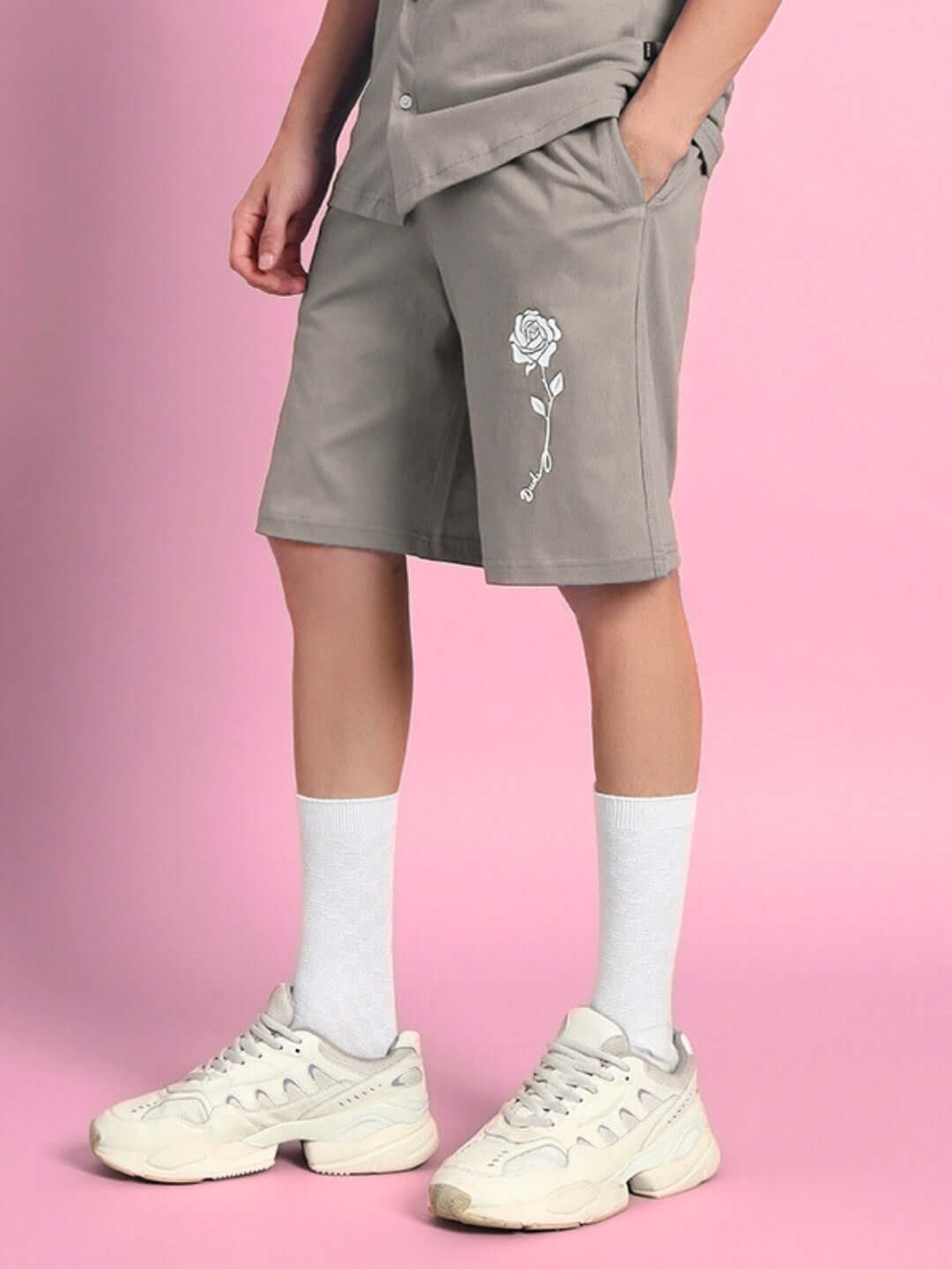 Person wearing LIMBO SUMMER COTTON SHORTS GREY with rose design, paired with white sneakers, against a pink background.