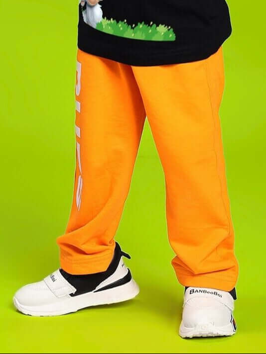 Child wearing LIMITED EDITION JOGGER FOR BOYS AND GIRLS ORANGE with white sneakers, standing against a bright green background.