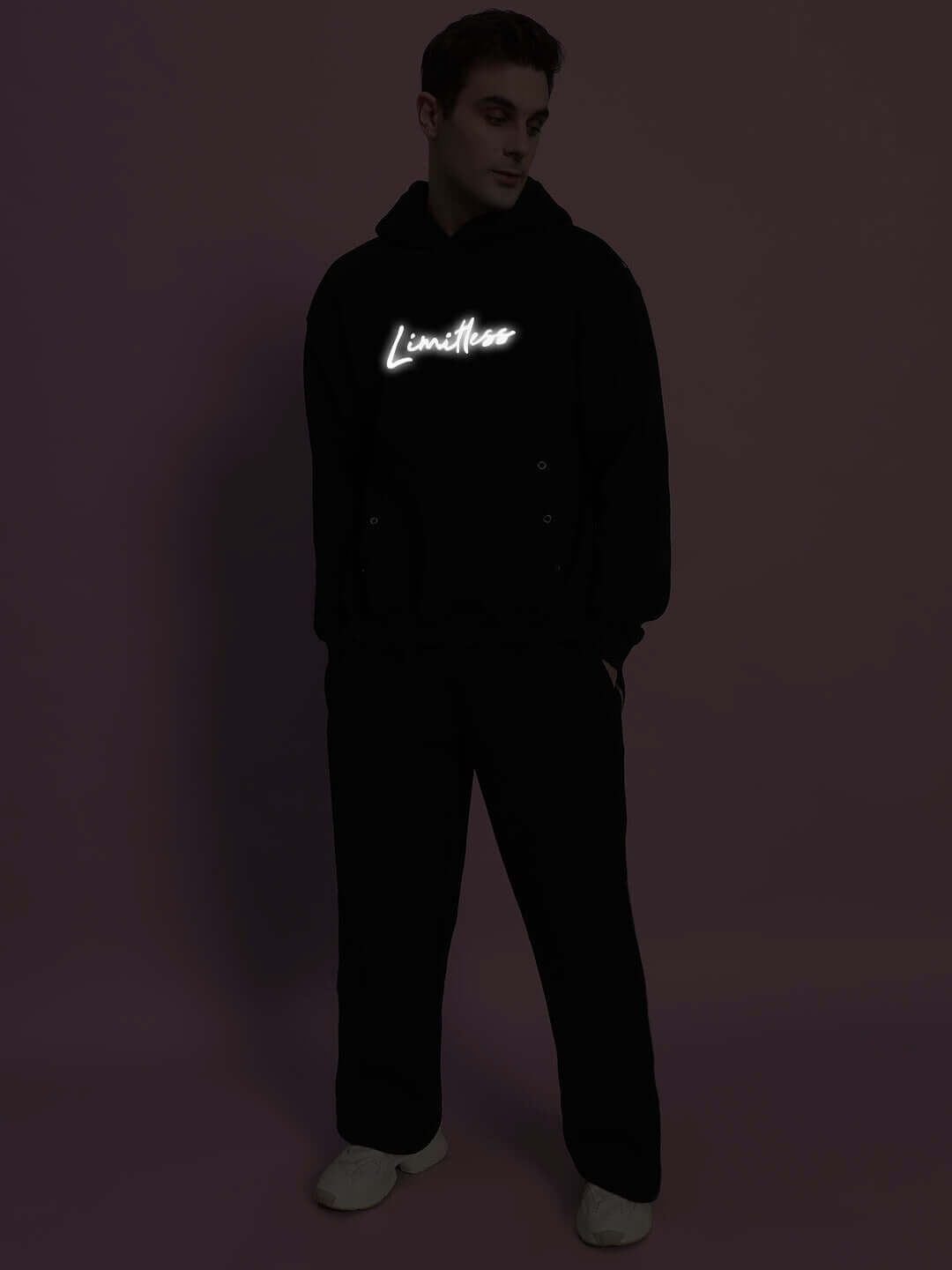 Model wearing LIMITLESS FLEECE REFLECTOR CO-ORD BLACK with glowing "Limitless" text on hoodie in dark setting.