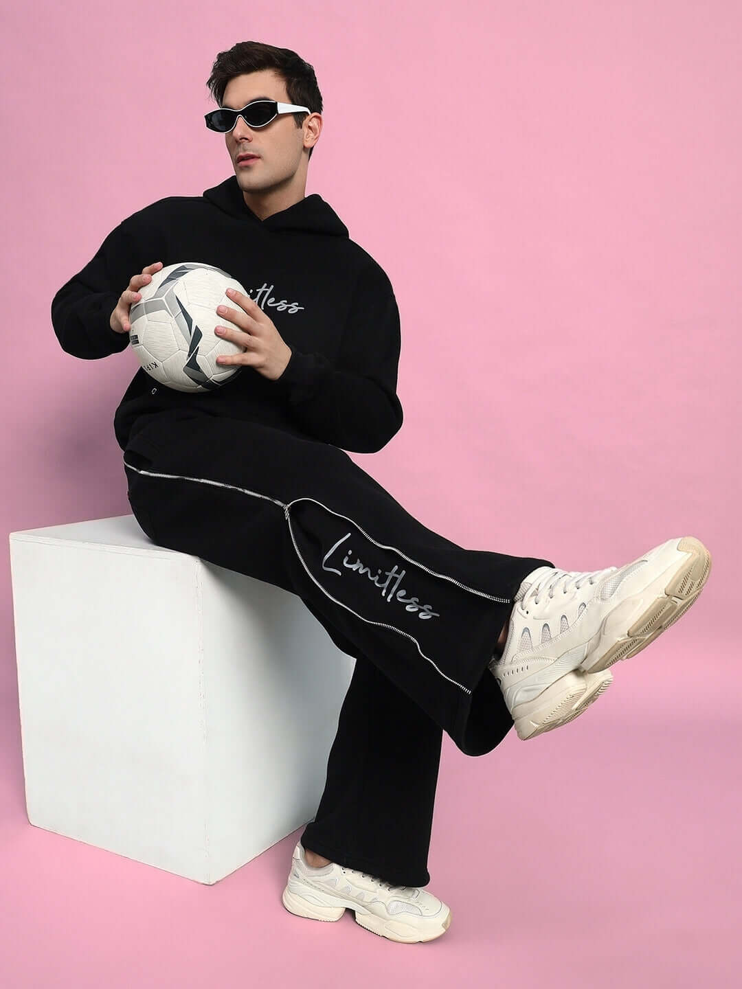 Model wearing LIMITLESS FLEECE REFLECTOR CO-ORD BLACK with sunglasses, holding a soccer ball against a pink background.