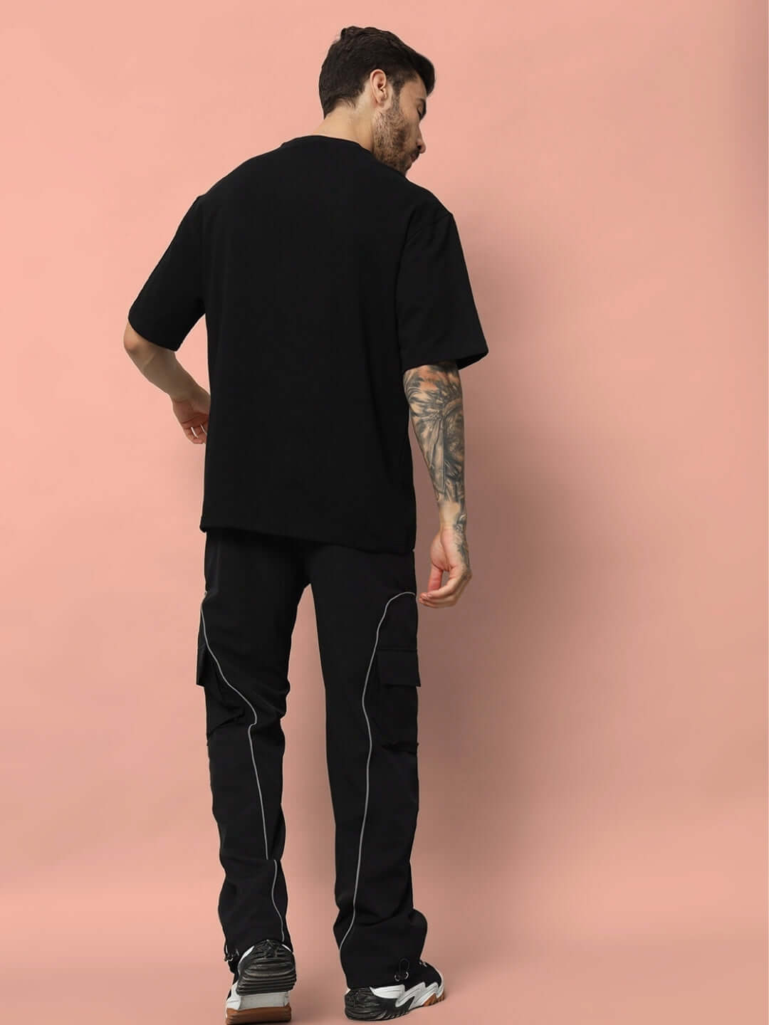 Man wearing LIMITLESS REFLECTOR OVER-SIZED T-SHIRT BLACK from back view with tattooed arm on a pink background.