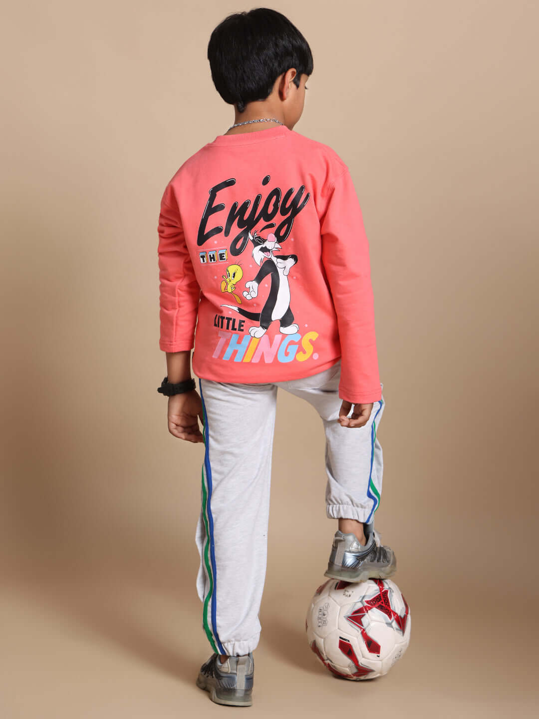 Young child wearing a peach and grey co-ord set with 'Enjoy the Little Things' text, standing with a soccer ball.