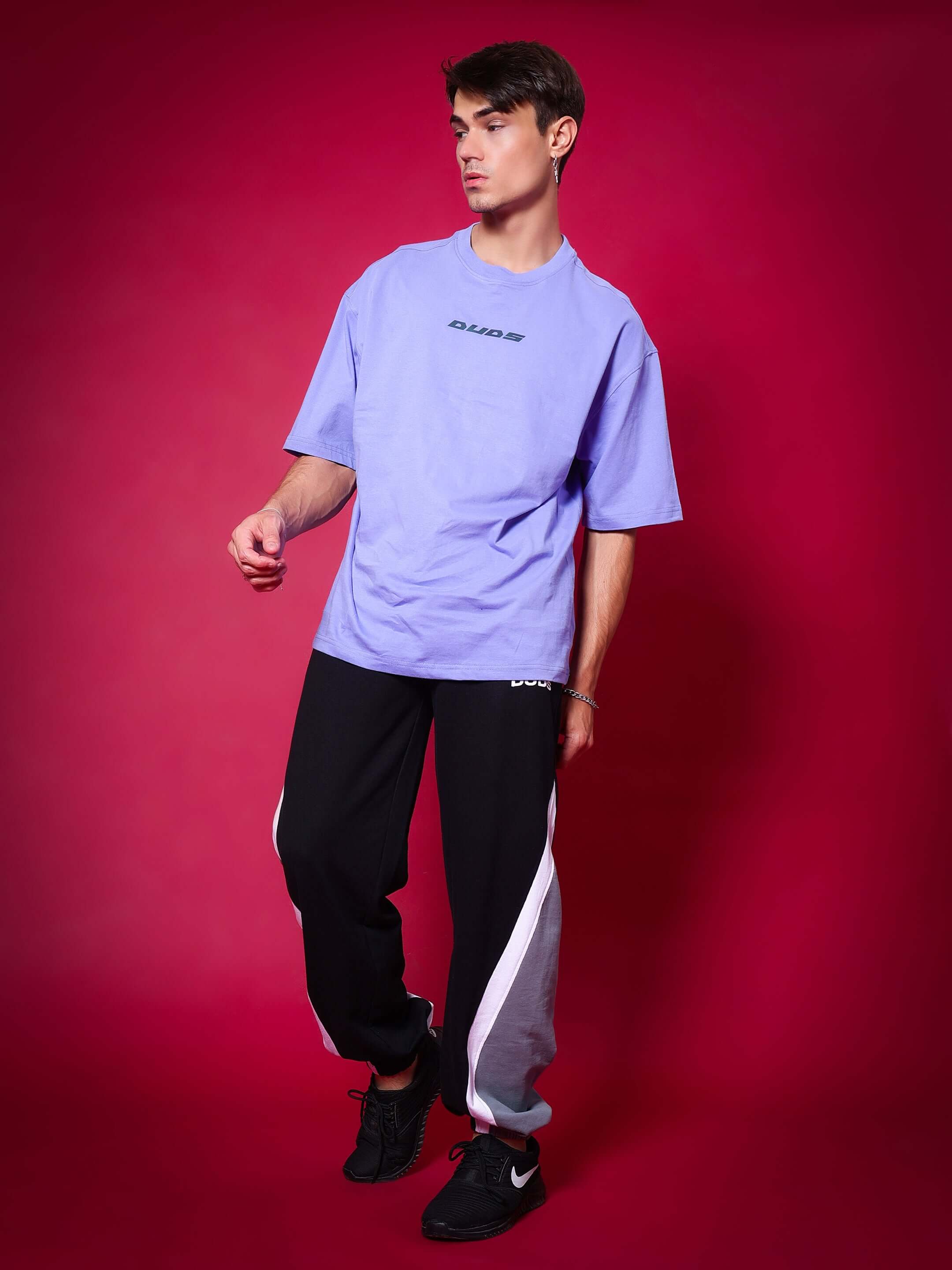 Model wearing Love Triangle Baggy Joggers in black-grey by DUDS, paired with purple shirt on a red background.