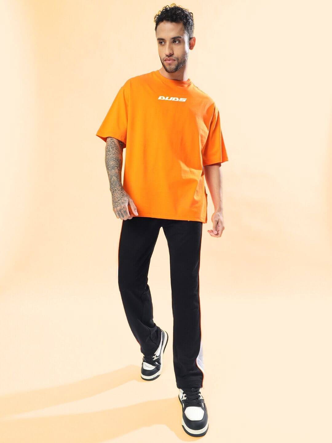 Man wearing a MACLERAN PRINTED OVERSIZED T-SHIRT ORANGE, standing against a light background with casual black pants and sneakers.