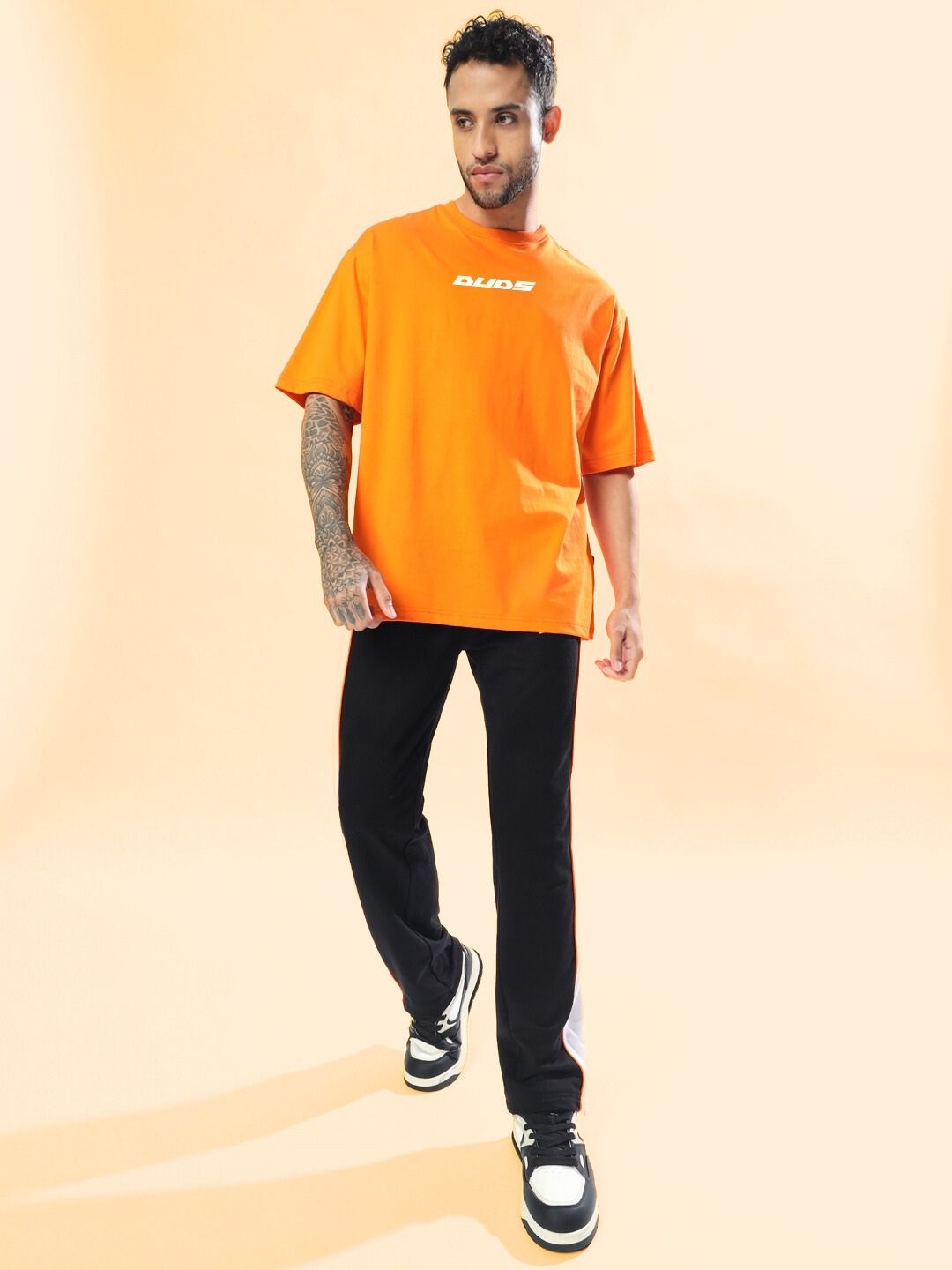 Man wearing MACLERAN PRINTED OVERSIZED T-SHIRT ORANGE with graphic design, paired with black pants, standing on a light background.
