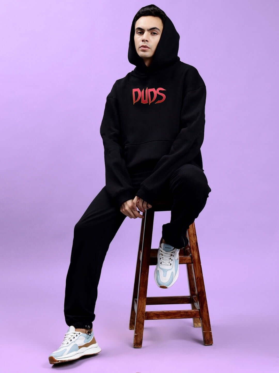 Model wearing a Mafia Fleece Oversize Hoodie in Black, sitting on a stool with a bold graphic design on the front.