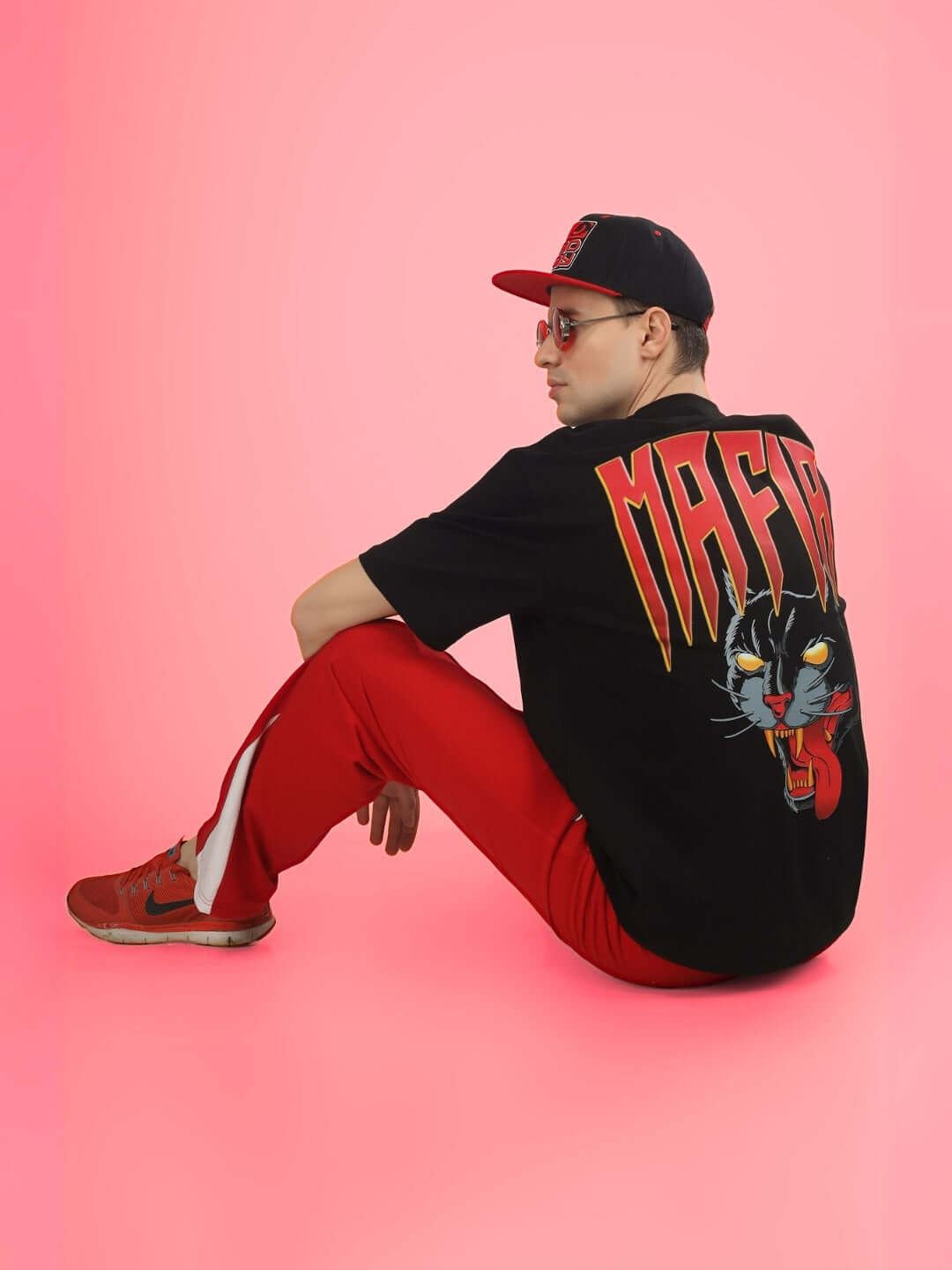 Person wearing Mafia oversized black T-shirt with graphic design, red pants, and a cap on a pink background.