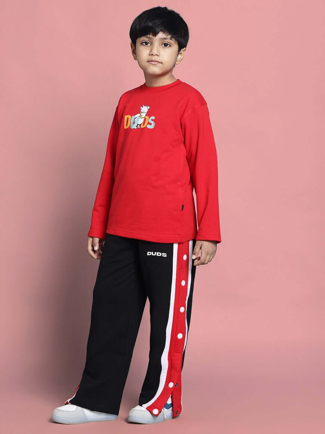 Young boy wearing red and black Winnie Co-ord set from DUDS, showcasing long-sleeve top and snap-button pants against a pink background.