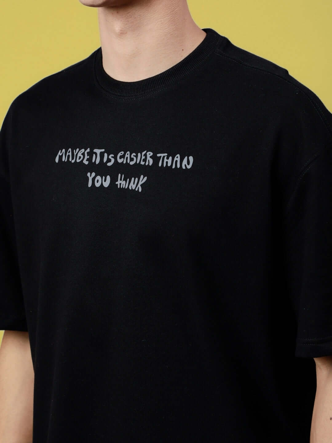 Black oversized T-shirt with 'MAYBE IT IS EASIER THAN YOU THINK' text in white, made from premium cotton blend, on yellow background.