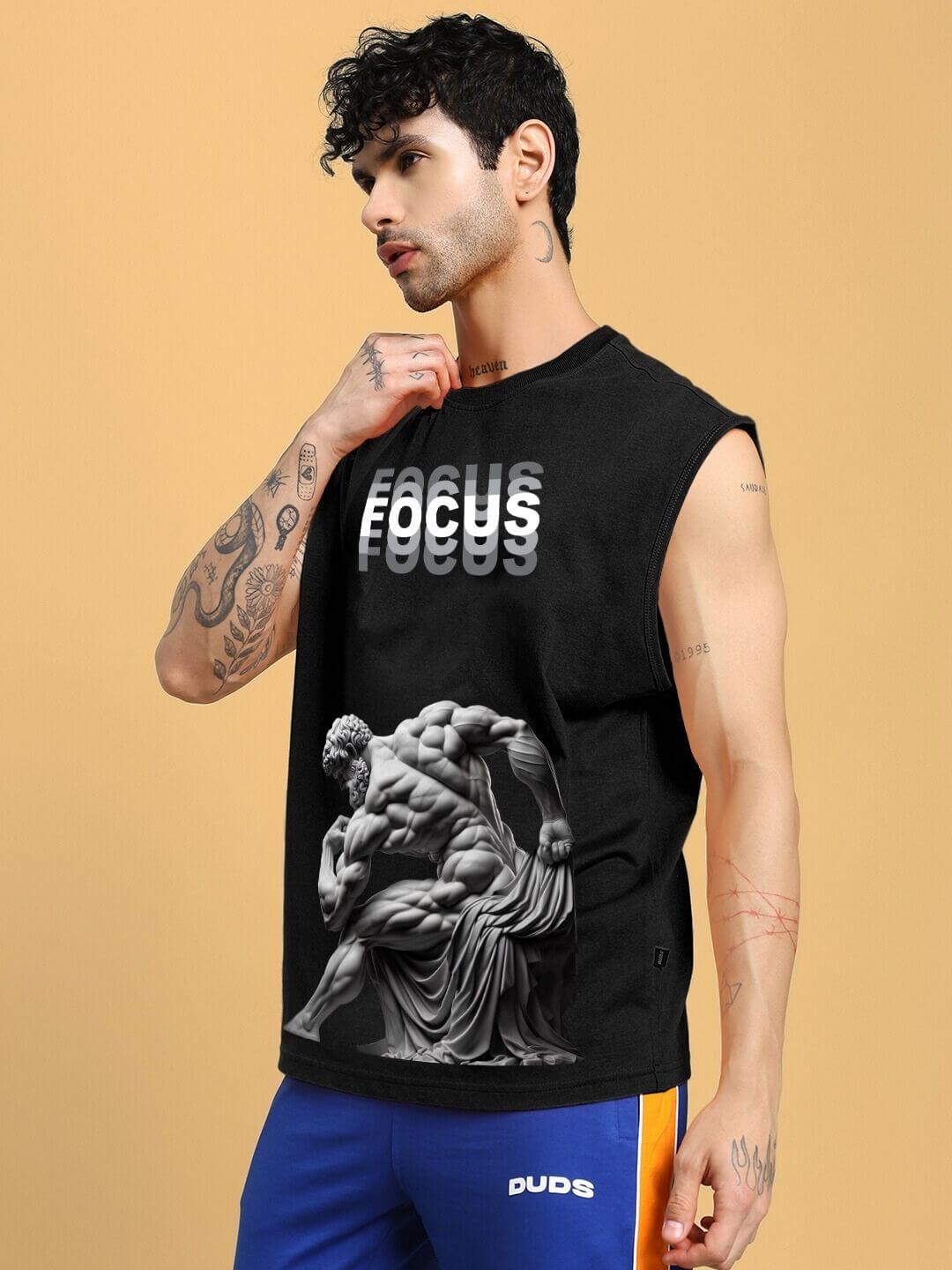 Man wearing JUST FOCUS FINE T-SHIRT in black, featuring artistic design, paired with blue and orange shorts, posing against a beige backdrop.