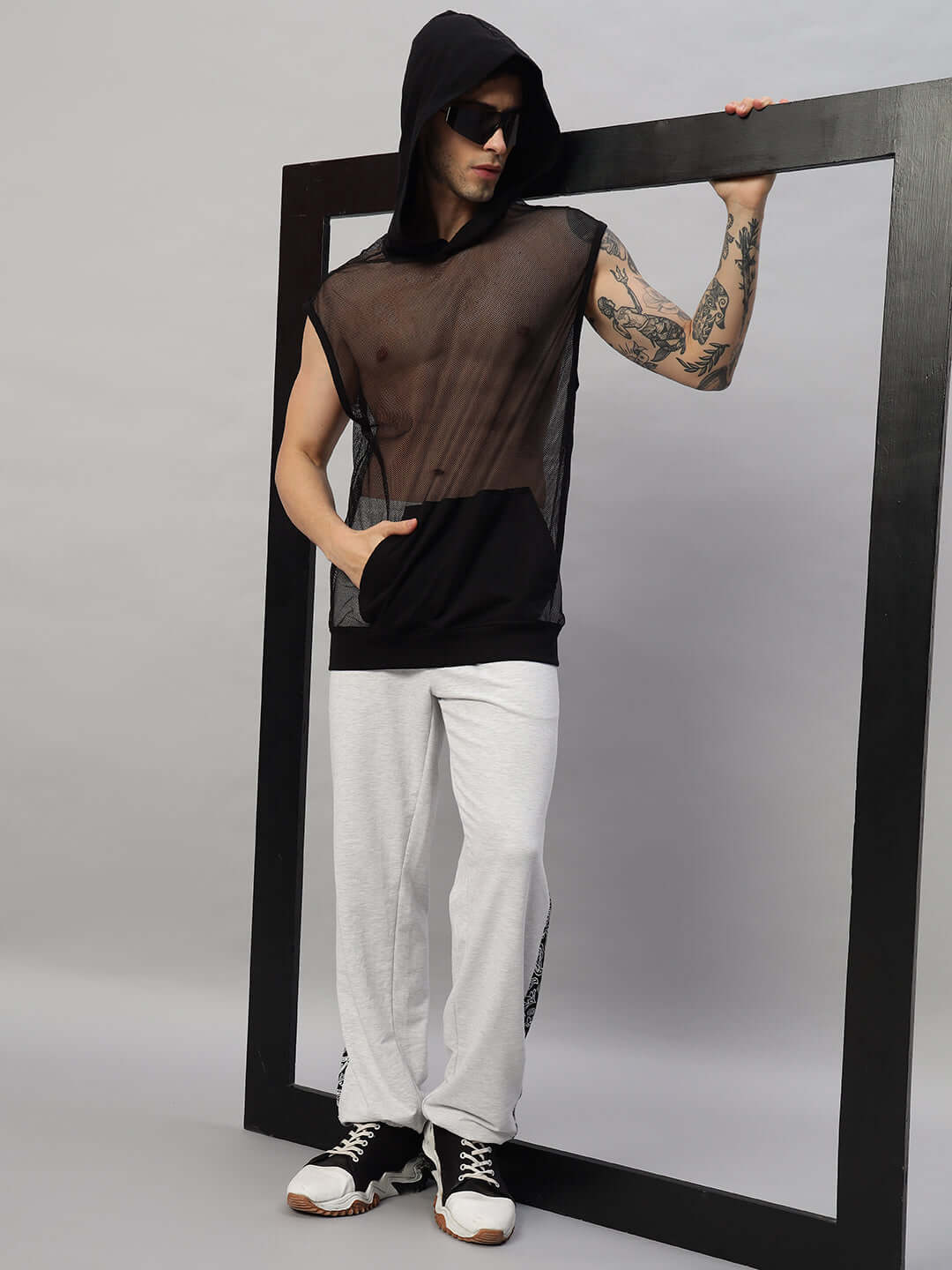 Model wearing stylish streetwear with a see-through sleeveless hoodie and white pants, leaning on a frame, fashion-forward look.