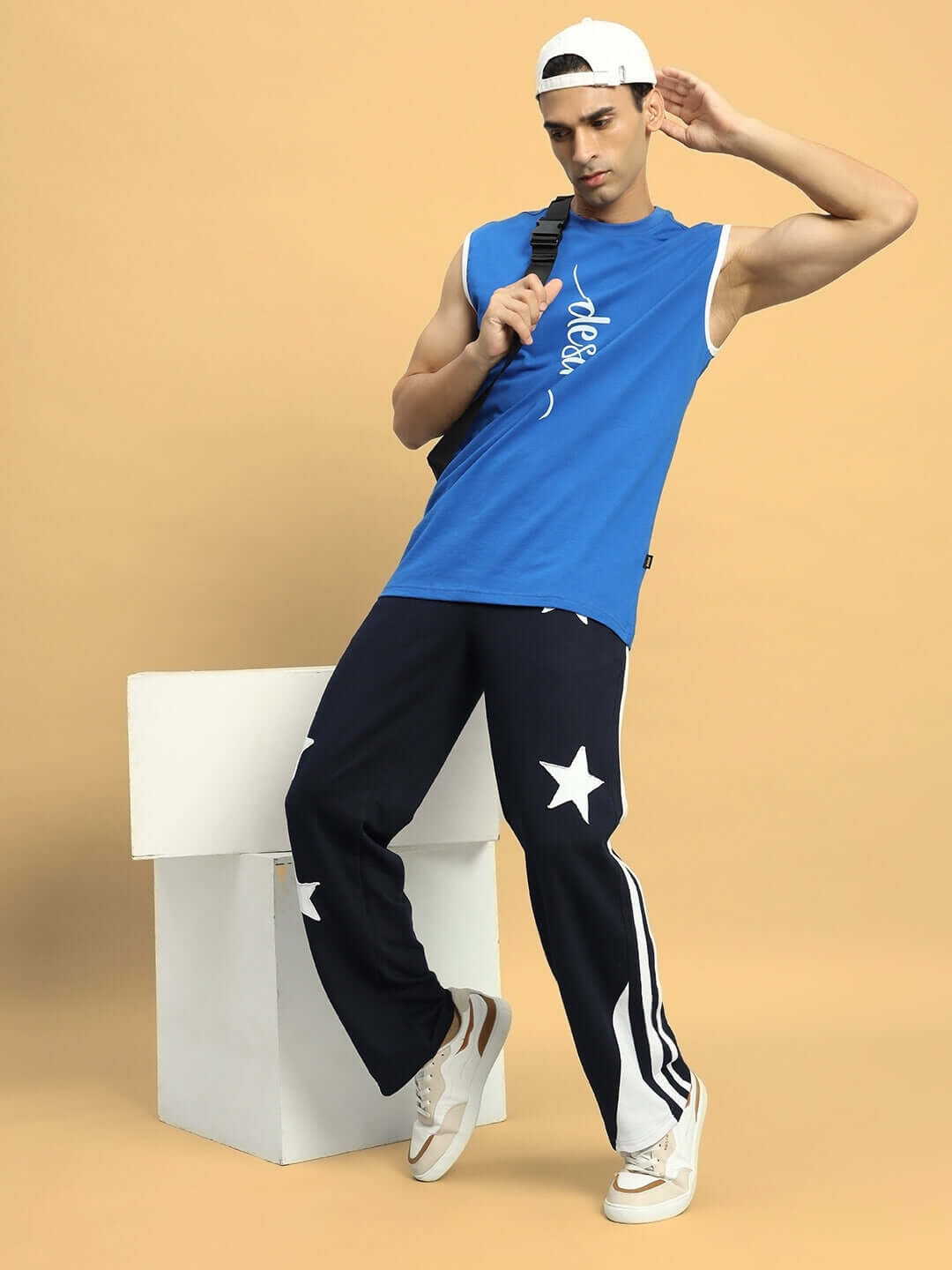 Man wearing navy blue DROOP RELAXED JOGGERS with star design, styled with a blue sleeveless shirt and white cap, posing against beige backdrop.