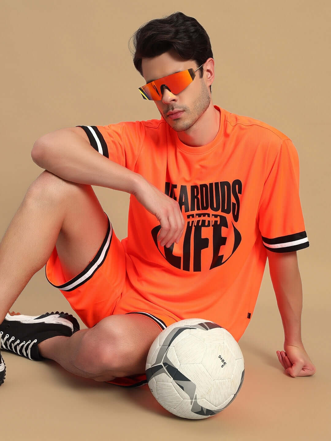 Alt Tag: Model wearing CHRONIC REGULAR FIT SHORTS ORANGE, sitting with a soccer ball, vibrant sportswear fashion, unisex design.