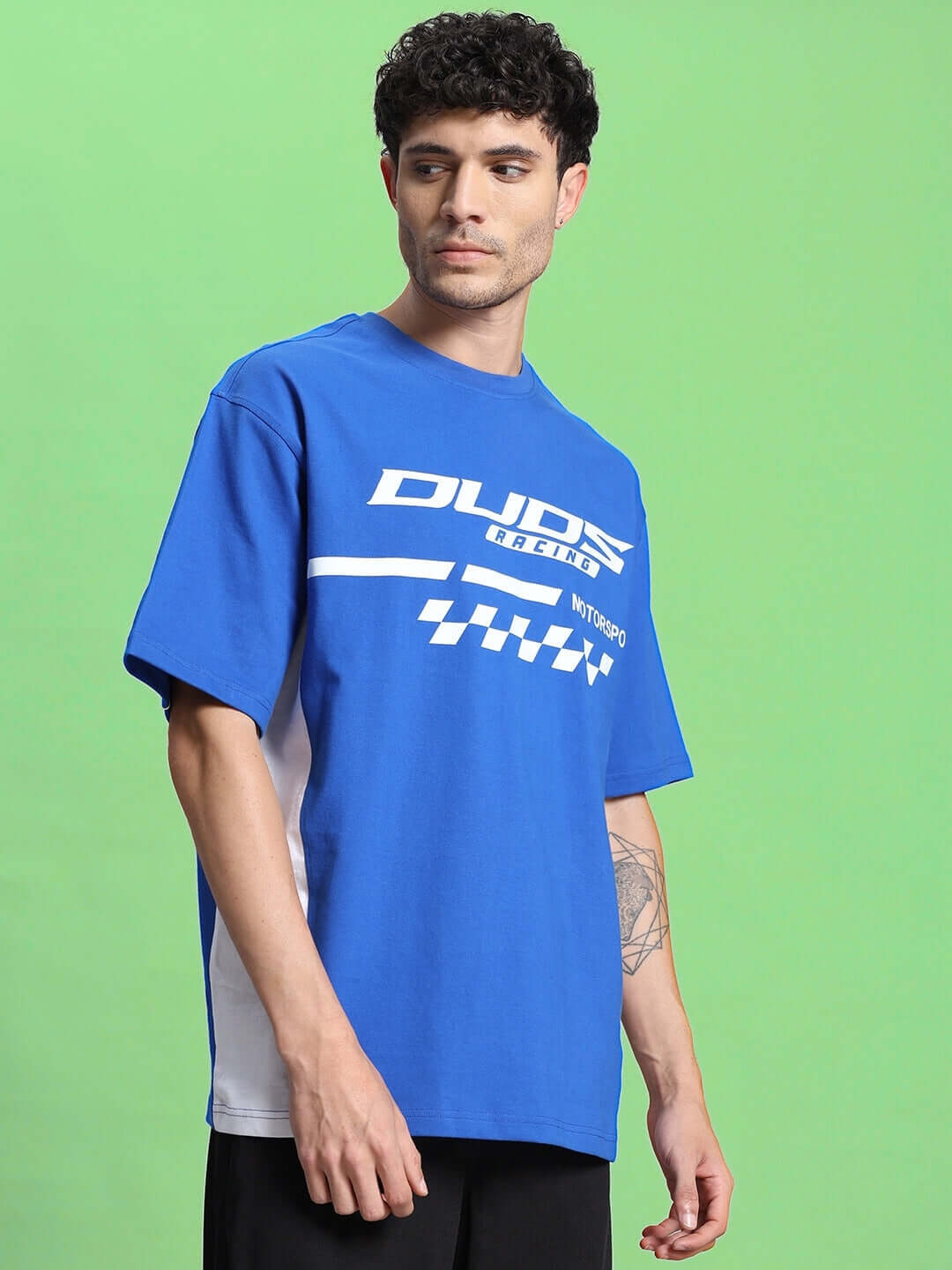 Man wearing INDY OVER-SIZED CLASSIC T-SHIRT ROYAL BLUE with streetwear design on green background.