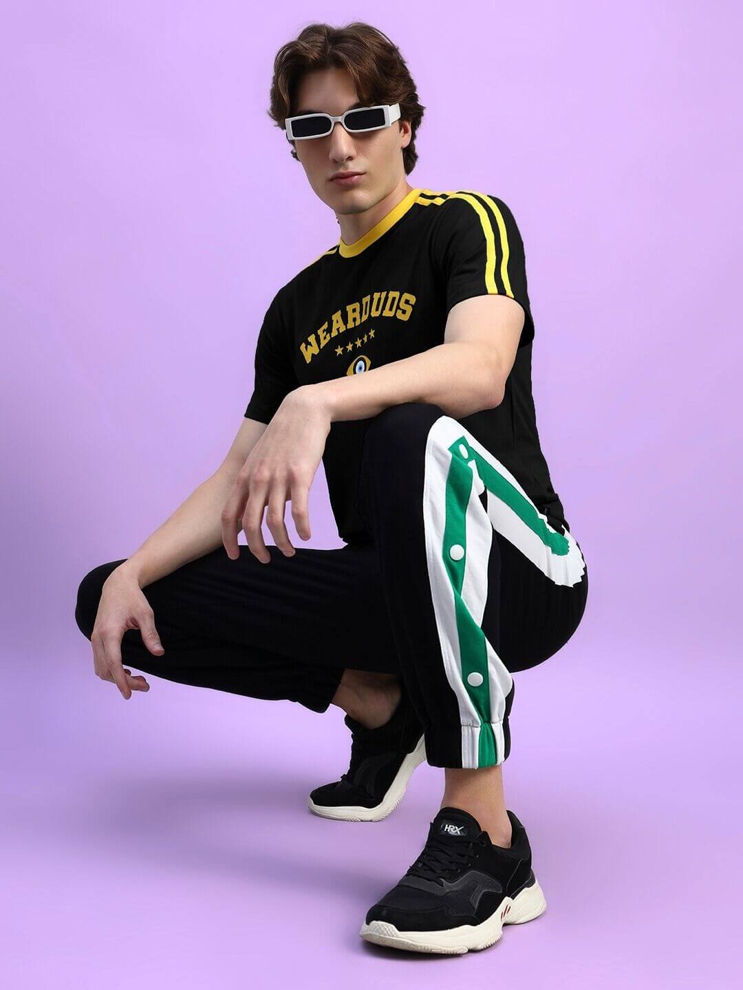 Man in sporty outfit including retro sunglasses and black sneakers, IRISH REGULAR FIT T-SHIRT BLACK,posing stylishly against a purple background.