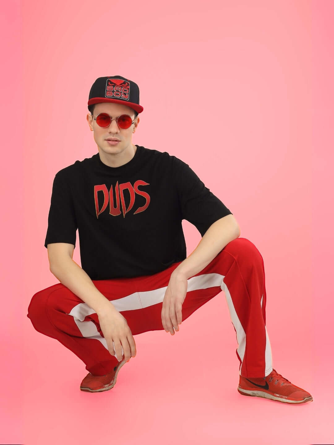 Model wearing DUDS Mafia oversized black t-shirt, red pants, and cap, crouching on pink background