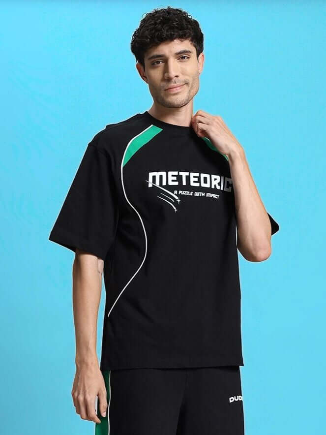 Model wearing METEORIC OVER-SIZED T-SHIRT BLACK in a relaxed pose against blue background, showcasing streetwear style and vibrant design.