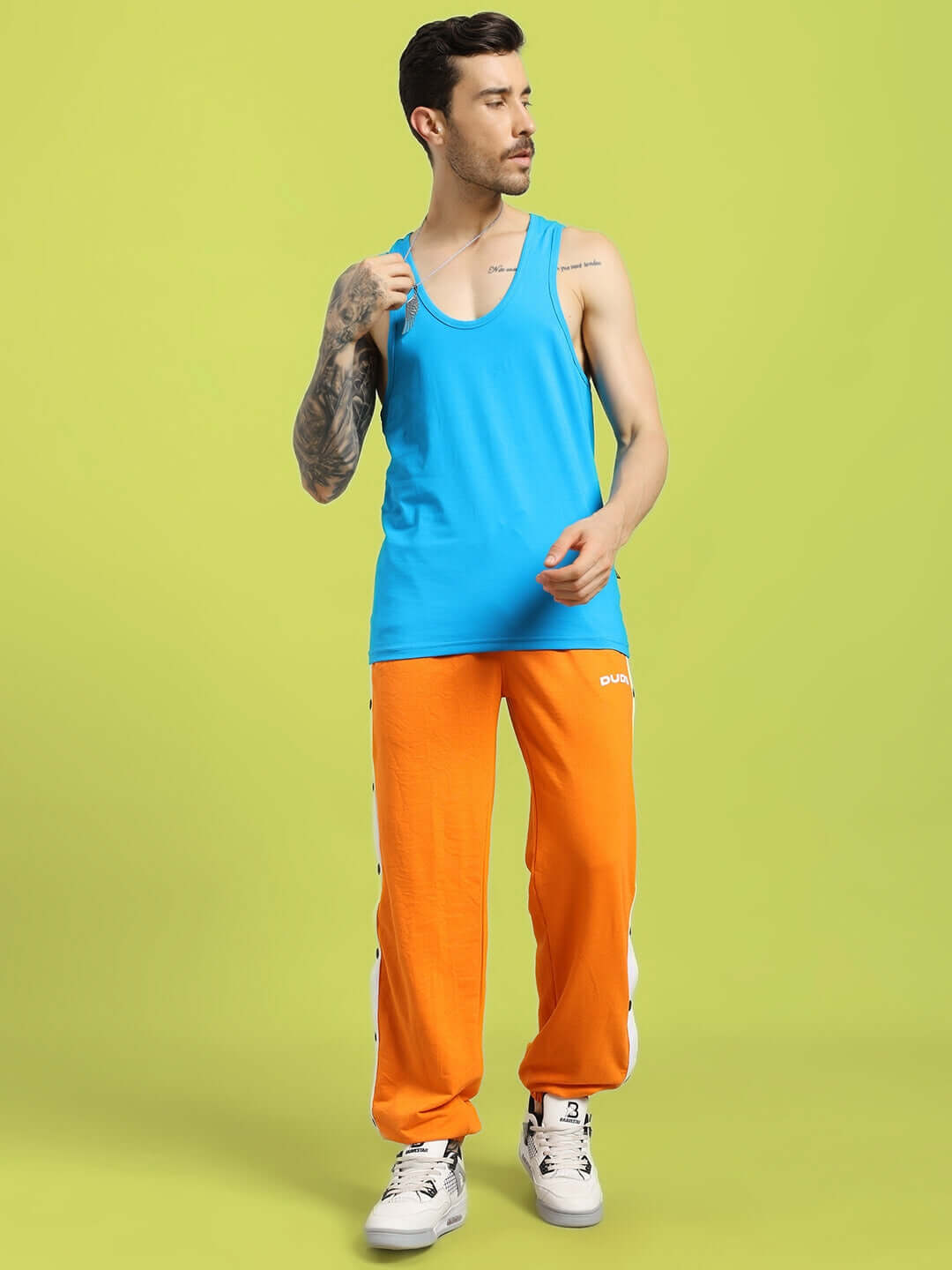 Man wearing Miller Relaxed Fit Joggers Orange paired with a blue tank top, showcasing a bold streetwear look on a green background.