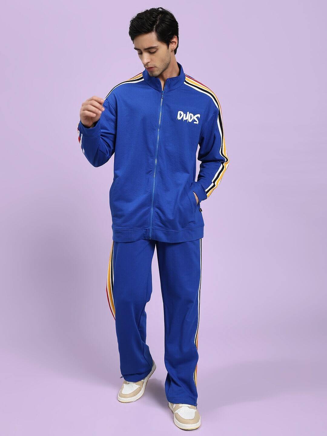 Man wearing the MODERN EDGE MULTI-STRIPE CO-ORD SET ROYAL BLUE with athletic style and colorful stripes, standing against a lavender background.