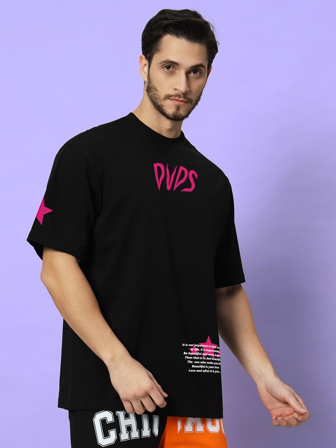 Male model wearing MODERN LOVE OVER-SIZED T-SHIRT BLACK with pink logo, standing against a purple background.
