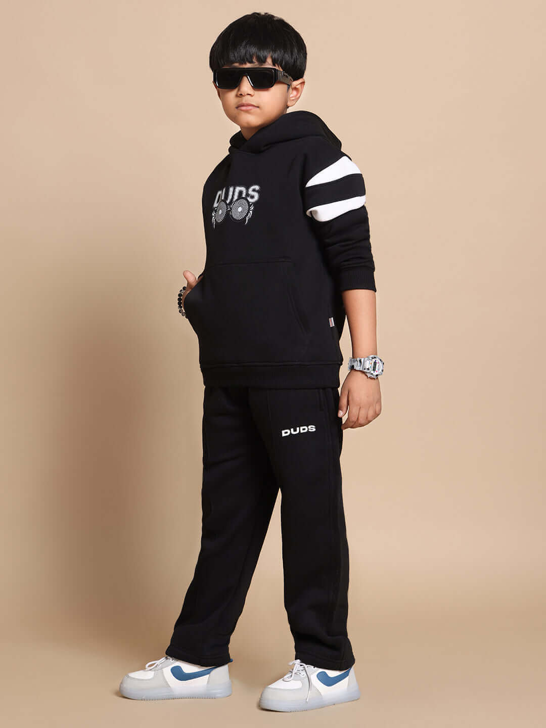 Child wearing a black Molly Jogger set for boys and girls by DUDS, featuring a hoodie and jogger pants, styled with sunglasses and sneakers.