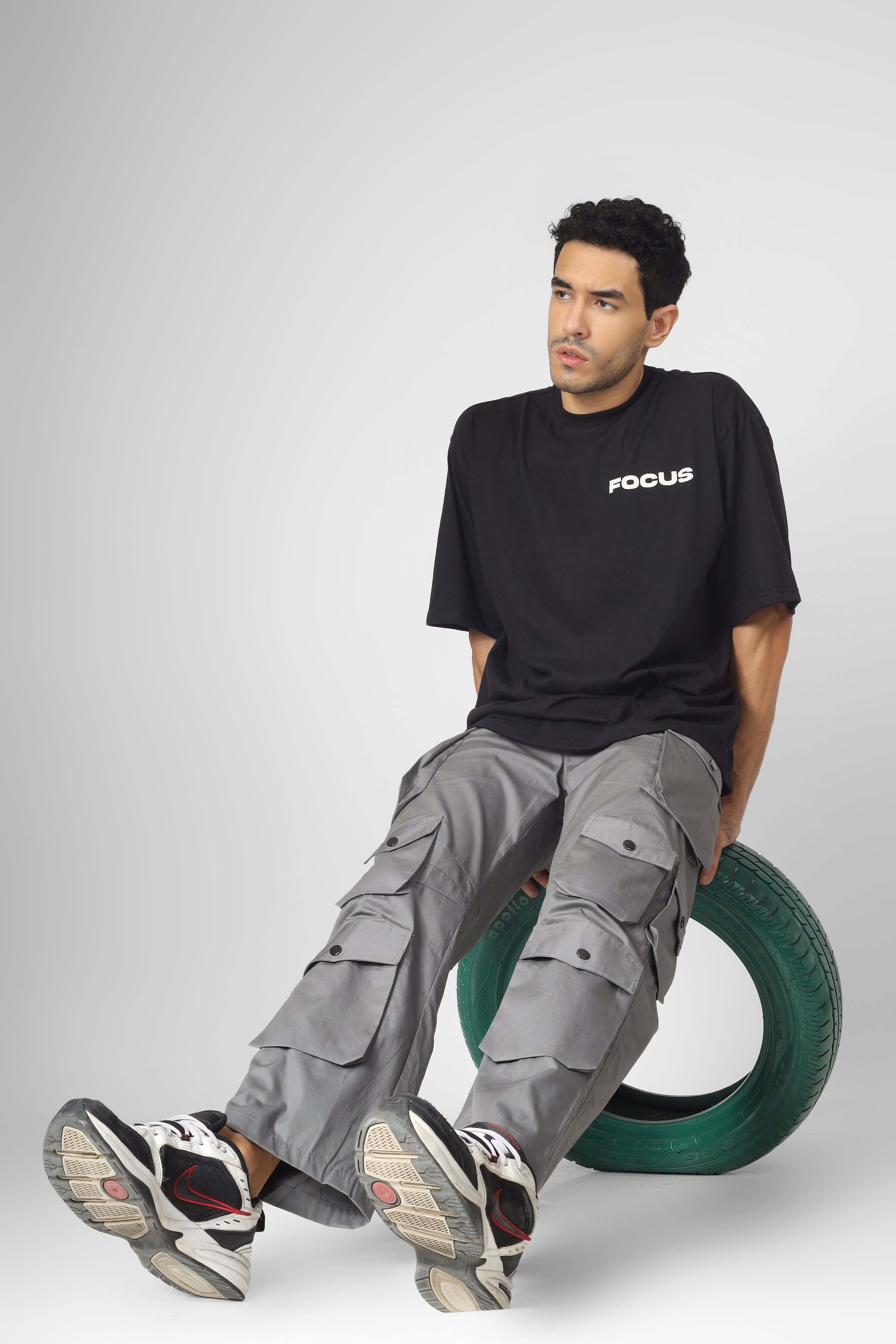 Model wearing MOON STONE cargo pants with 12 pockets, sitting on a tire, pairing the pants with a black t-shirt and sneakers.