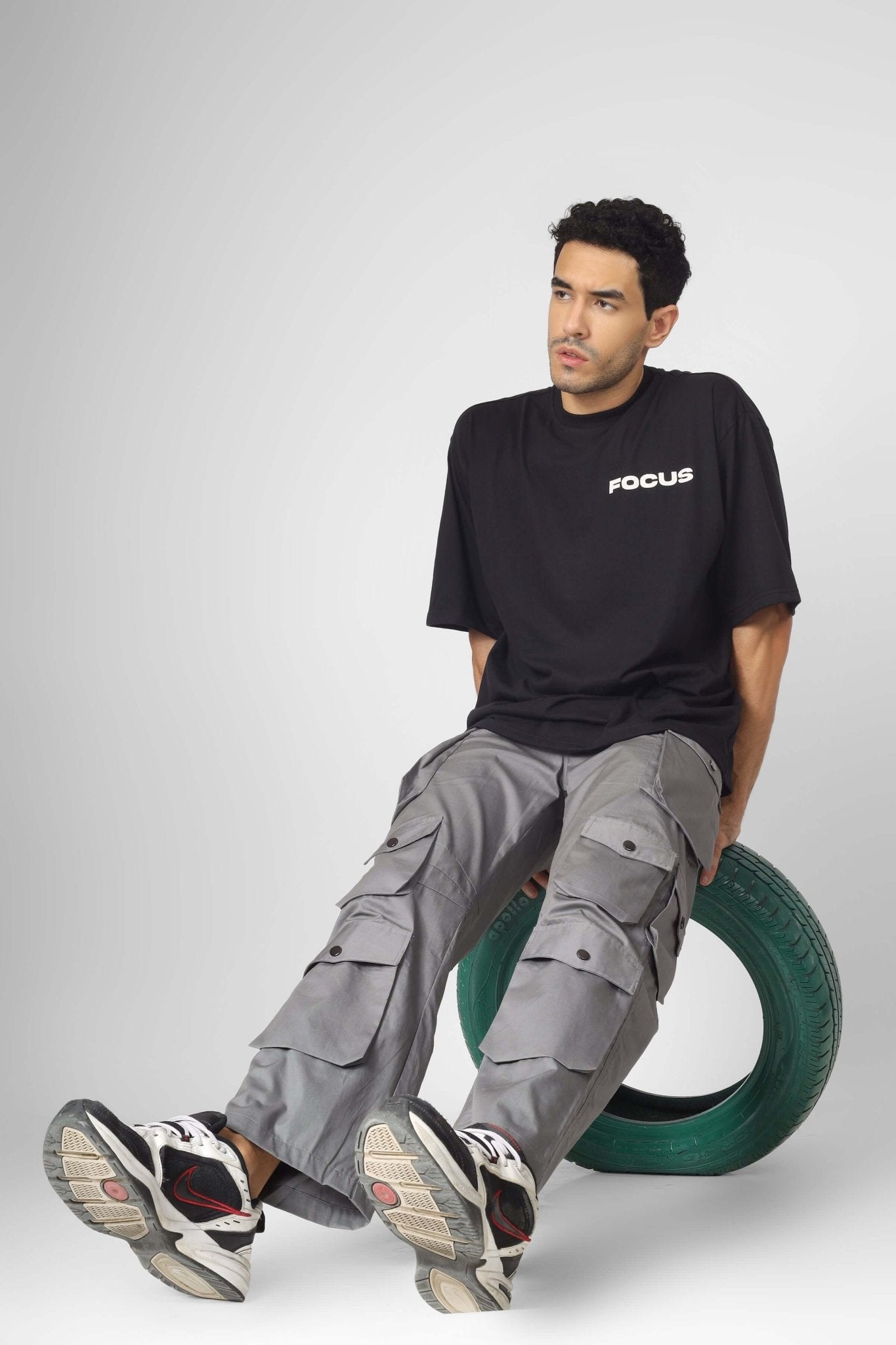 Man wearing MOON STONE CARGO PANTS WITH 12 CARGO POCKET sitting on a tire, showcasing urban streetwear style.