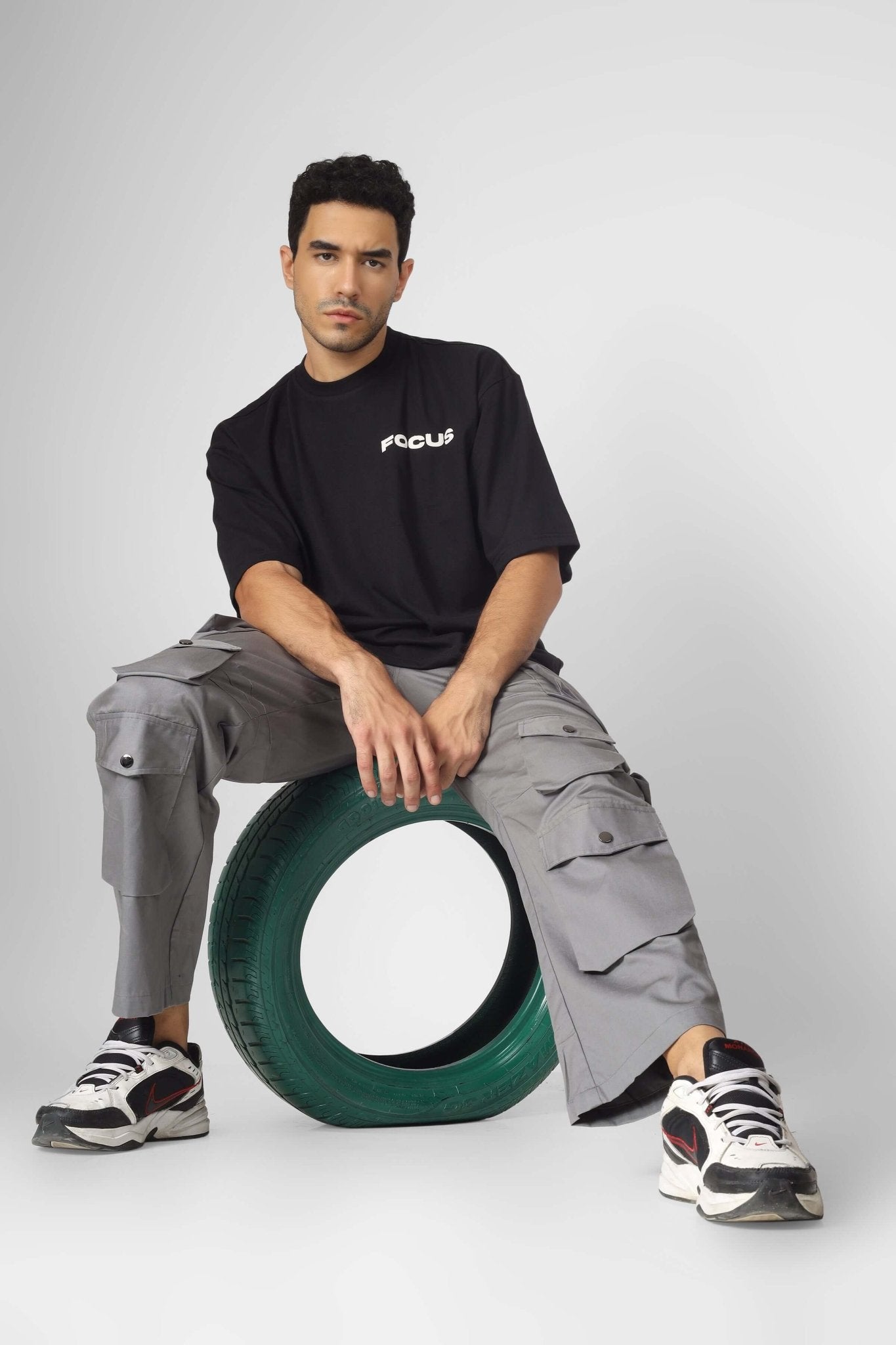 Man wearing MOON STONE CARGO PANTS WITH 12 CARGO POCKETS sitting on a tire, showcasing urban streetwear style and functionality.
